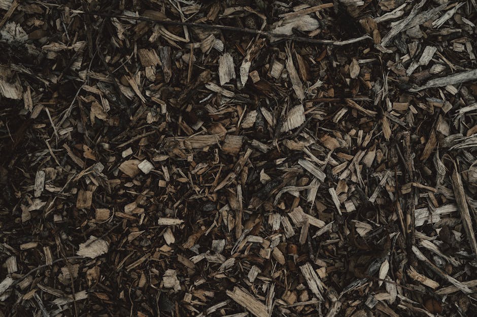 Close-up view of a pile of wood chip mulch with small, irregularly shaped pieces of bark and shredded wood, showcasing a rough texture with shades of brown and dark gray. The mulch appears freshly chi